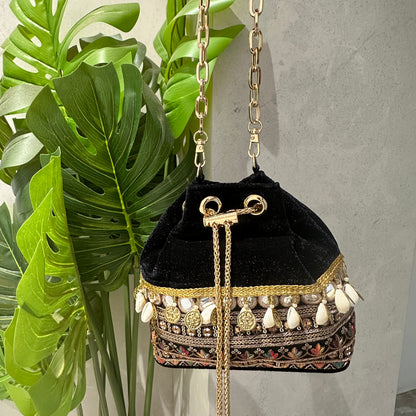 A black velvet bucket bag with multicolor thread embroidery, gold coin charms, and a gold-toned chain handle. It is displayed next to a green plant.