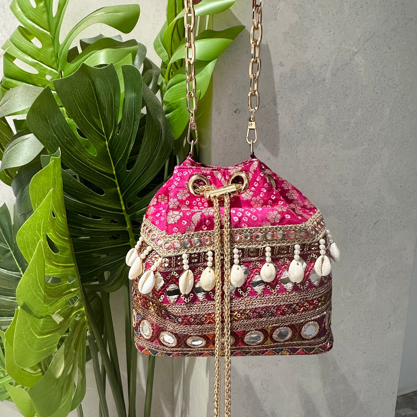 A handcrafted pink bandhani fabric bucket bag with multicolored thread embroidery, mirror work, cowrie shells, and antique gold coin charms. It features a gold-toned chain handle and drawstring closure with golden tassels.