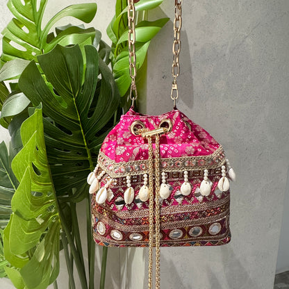 A handcrafted pink bandhani fabric bucket bag with multicolored thread embroidery, mirror work, cowrie shells, and antique gold coin charms. It features a gold-toned chain handle and drawstring closure with golden tassels.